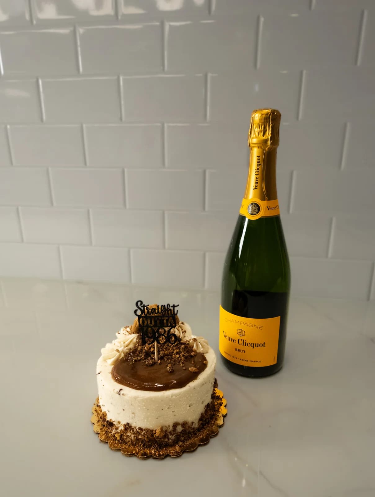 Birthday cake with caramel drizzle and a "Straight Outta 1986" topper beside a bottle of Veuve Clicquot champagne on a white marble counter.