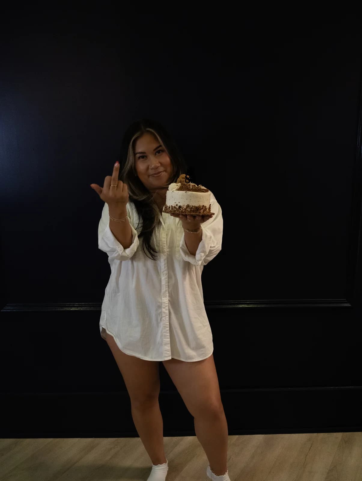 Woman making a playful middle-finger gesture with a smirk while holding a birthday cake in front of a navy wall.