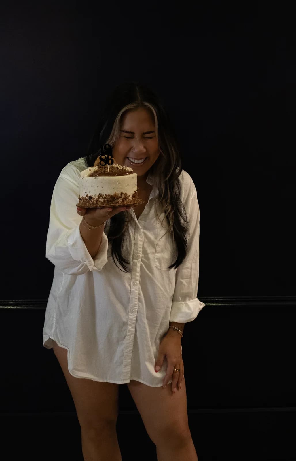 Woman laughing with eyes closed behind a birthday cake held close to her face, wearing a loose white shirt.