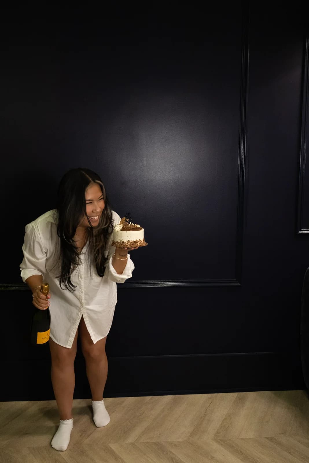 Woman laughing as she leans forward with a birthday cake in one hand and a champagne bottle lowered at her side.