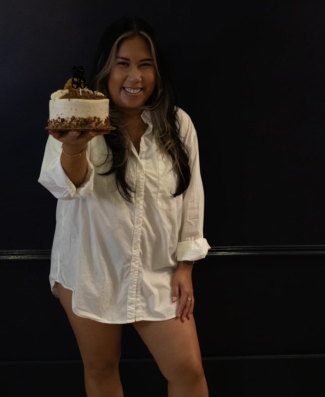 Woman smiling broadly while holding a birthday cake out toward the camera with her hand resting on her leg.