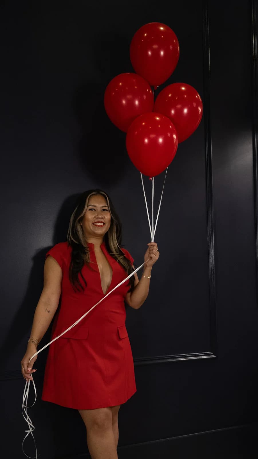 Close, smiling portrait of a woman in a red blazer dress holding a bunch of glossy red balloons.