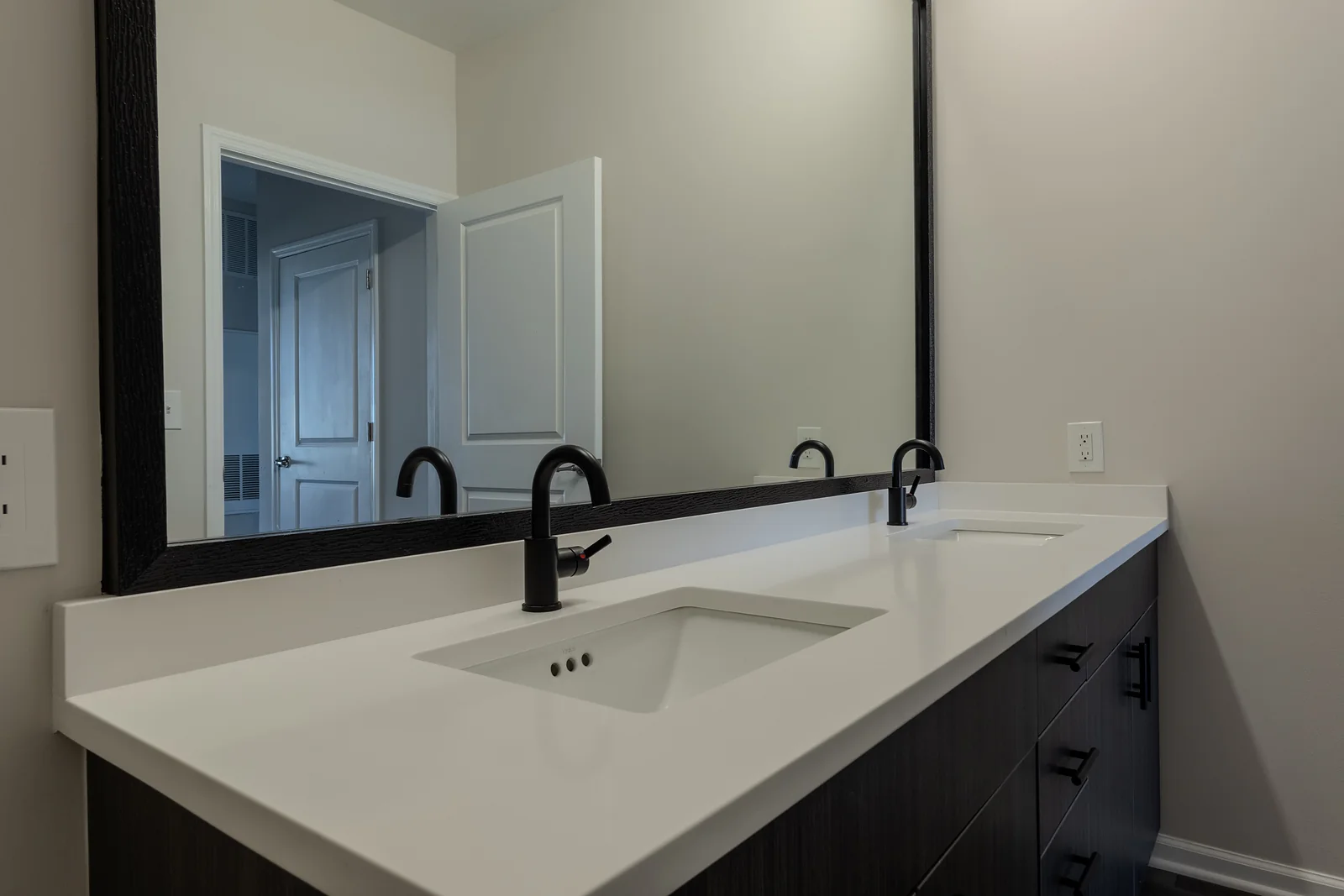 White bathroom countertop with an undermount sink and matte black faucets beneath a black-framed mirror.