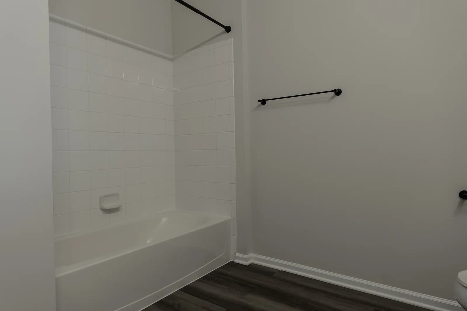 Bathroom with a white tiled tub and shower, matte black curtain rod and towel bar, and gray plank floor.