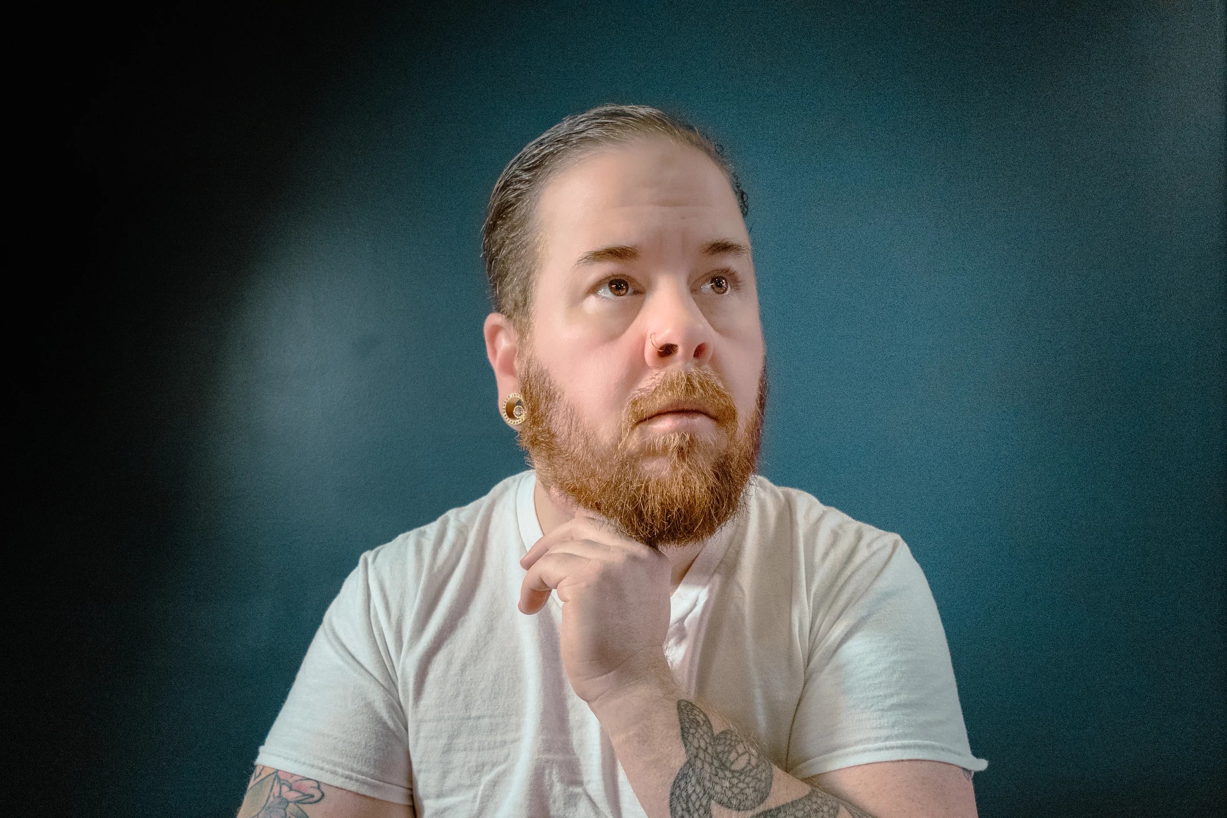 Self-portrait of photographer Andrew Liam with a red beard and slicked-back hair, resting his chin on his hand and gazing upward against a dark teal backdrop.