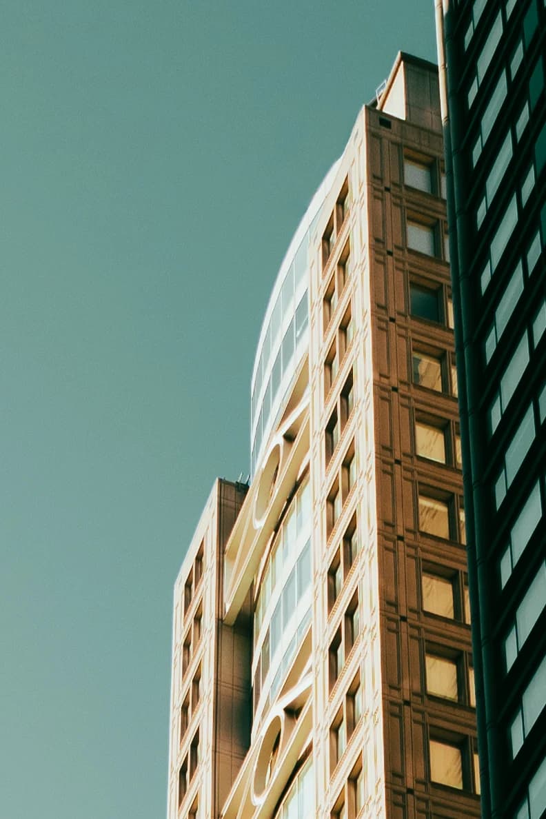 Color photo looking up at a tall building in New York City. Late-afternoon sun lights a warm stone facade with a grid of square windows and a curved glass section with oval cutouts, set against a clear teal sky, with a dark glass tower along the right edge.