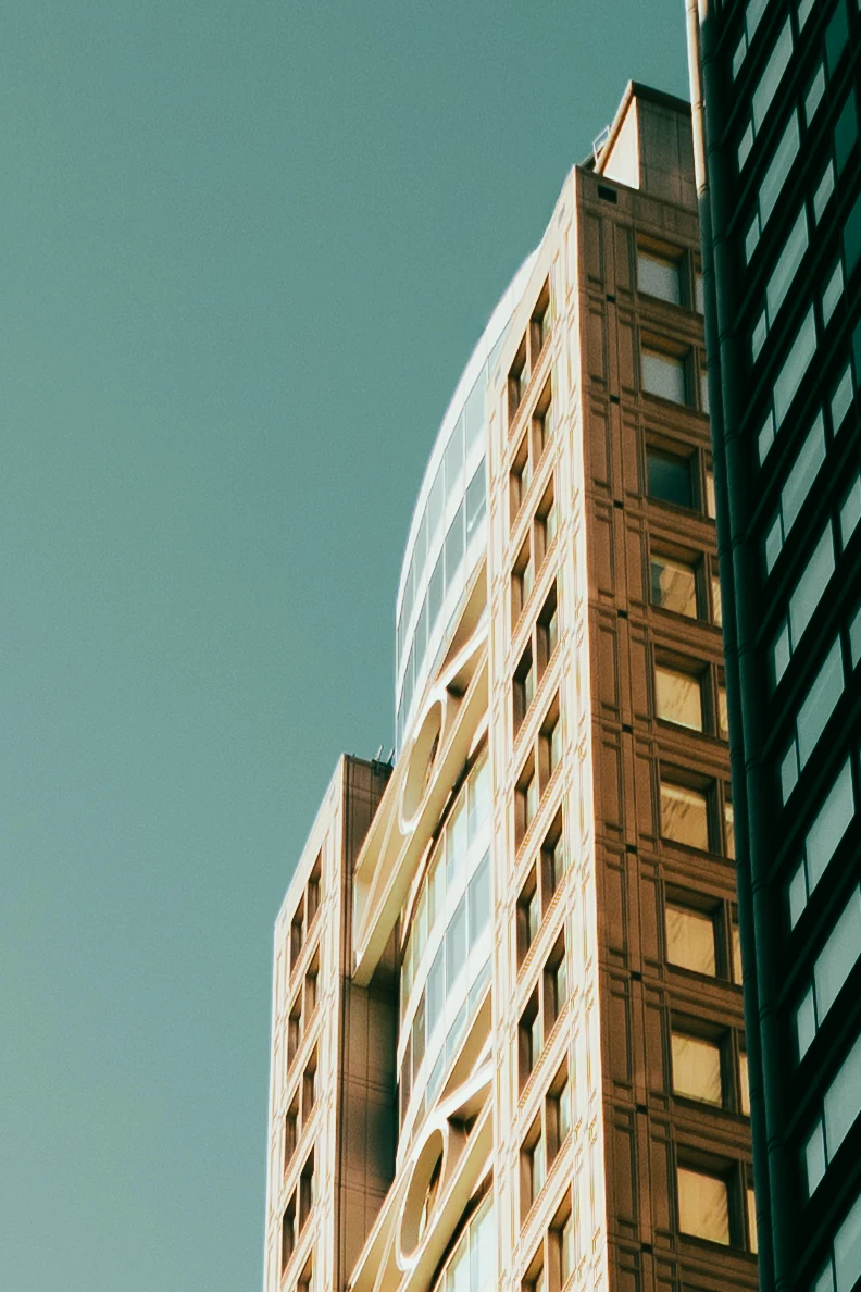 Color photo looking up at a tall building in New York City. Late-afternoon sun lights a warm stone facade with a grid of square windows and a curved glass section with oval cutouts, set against a clear teal sky, with a dark glass tower along the right edge.