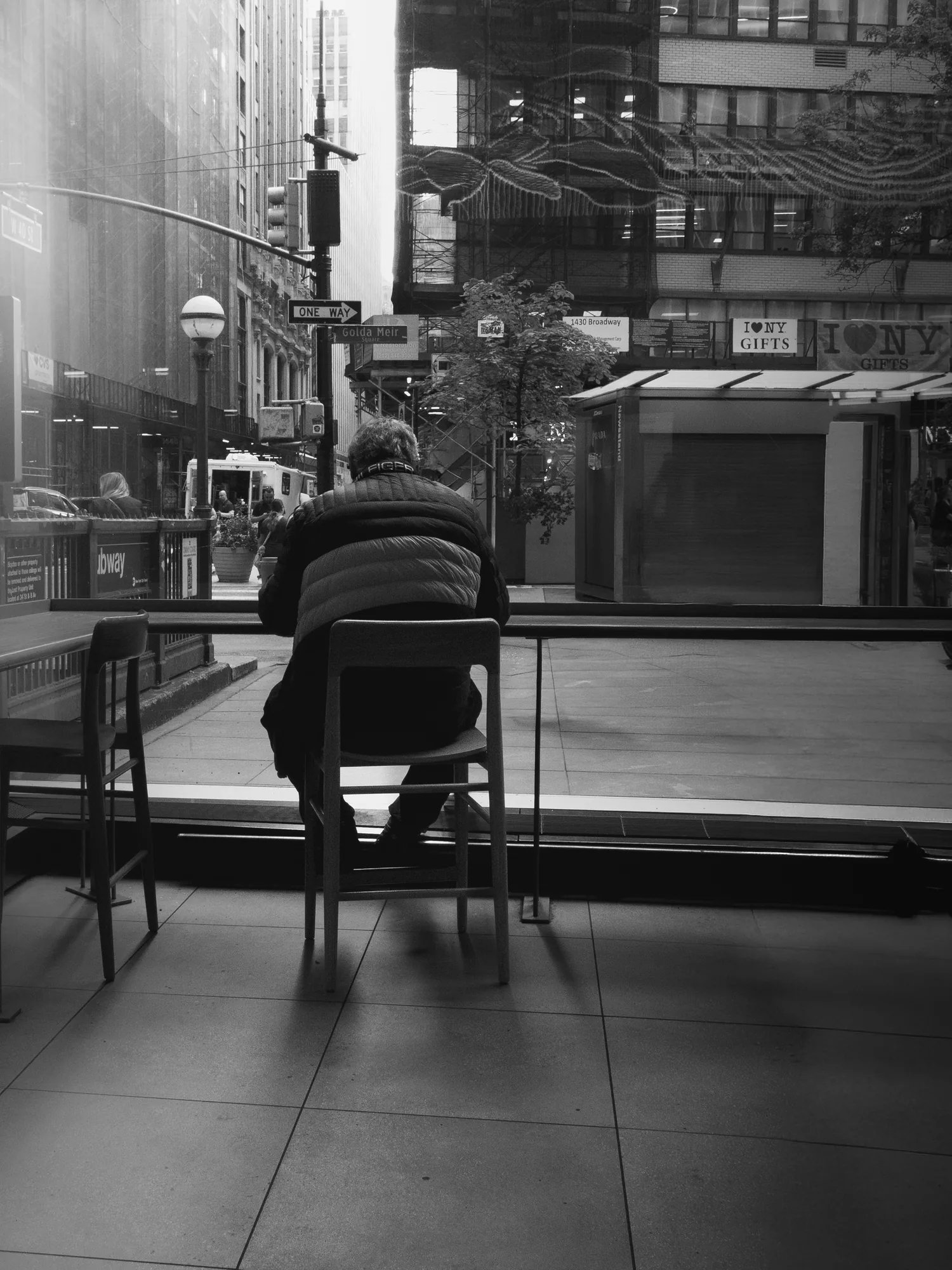 Black-and-white photo taken from inside a window in Midtown Manhattan. A man in a quilted puffer jacket sits on a tall wooden stool with his back to the camera, looking out at Golda Meir Square, where a One Way sign, a globe streetlamp, a subway entrance, and a shuttered newsstand under I Love NY Gifts signs line the corner.