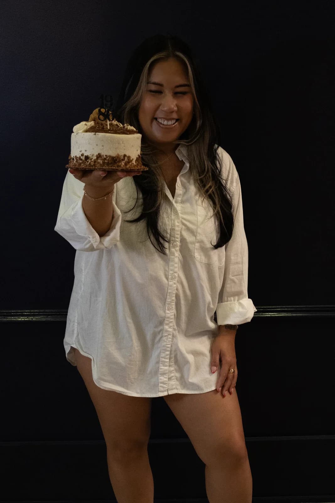 Woman grinning and presenting a frosted birthday cake toward the camera in a white oversized shirt.